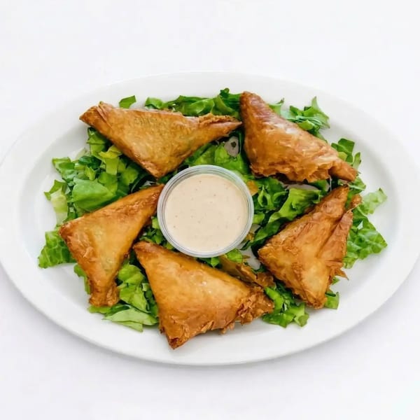 Five golden-brown triangular pastries, possibly samosas or spanakopita, arranged on a bed of fresh green lettuce with a small cup of dipping sauce in the center.